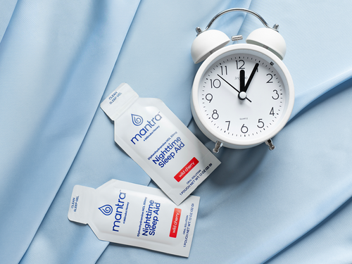 Two white Mantra Nighttime Sleep Aid pouches arranged next to a white twin-bell alarm clock on a light blue fabric background.